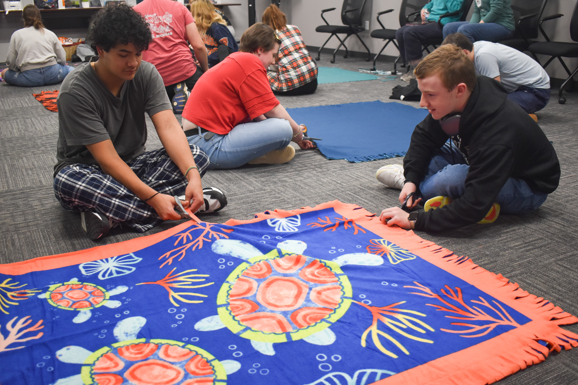Two TEAM members sitting on the ground making no-sew blankets.