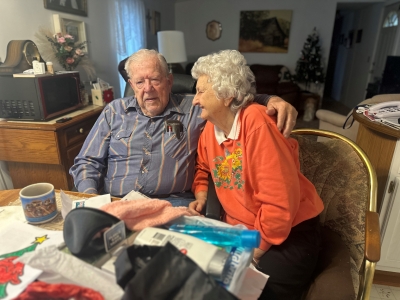 An older couple sitting down at a kitchen table. The man has his arm outstretched toward his wife. 