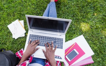 An overhead view of a person sitting in the grass with a laptop on their lap and notebooks scattered around them.