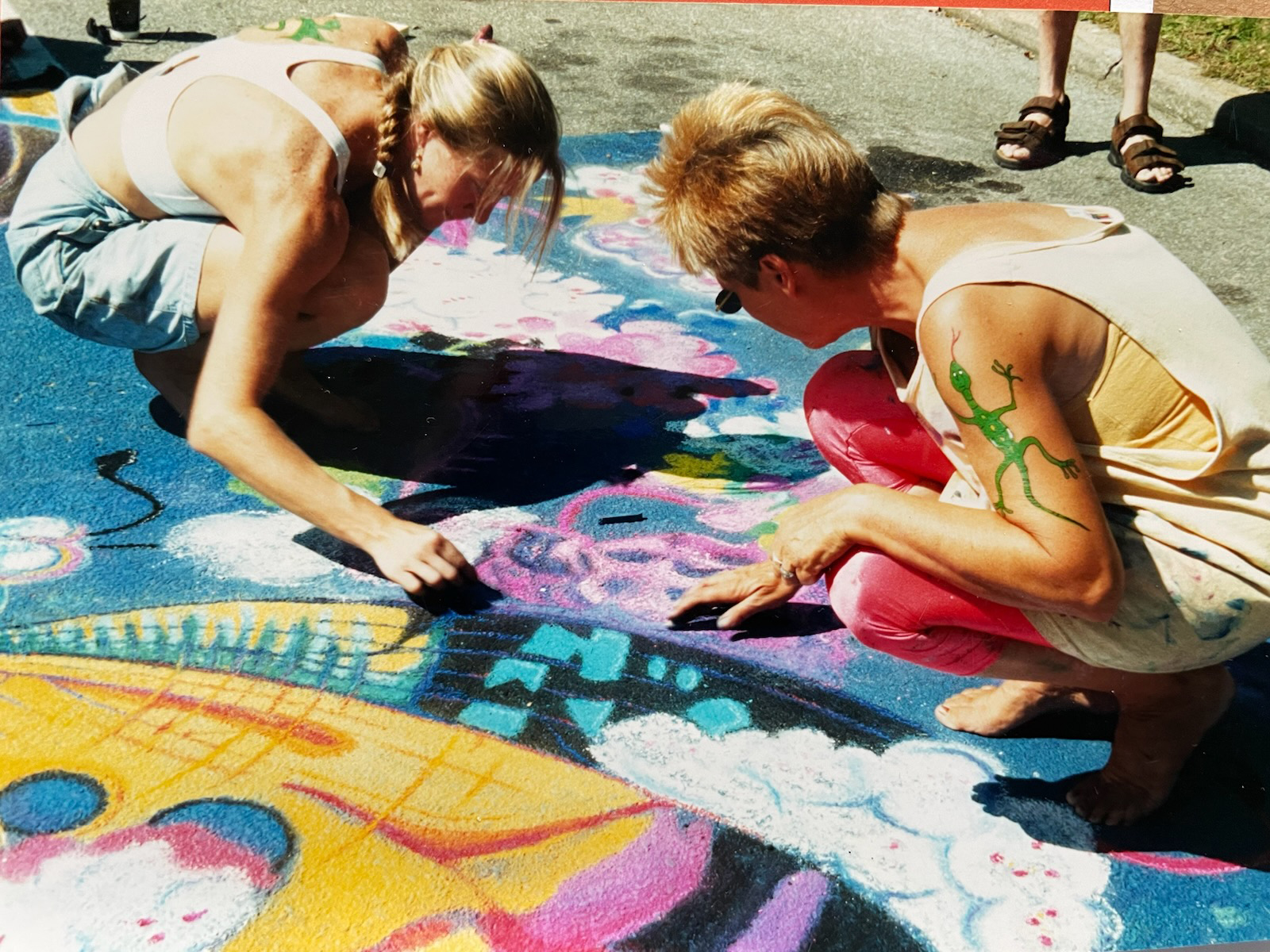 Nicole, left, and Judy are crouched close to the cement making vibrant chalk art.